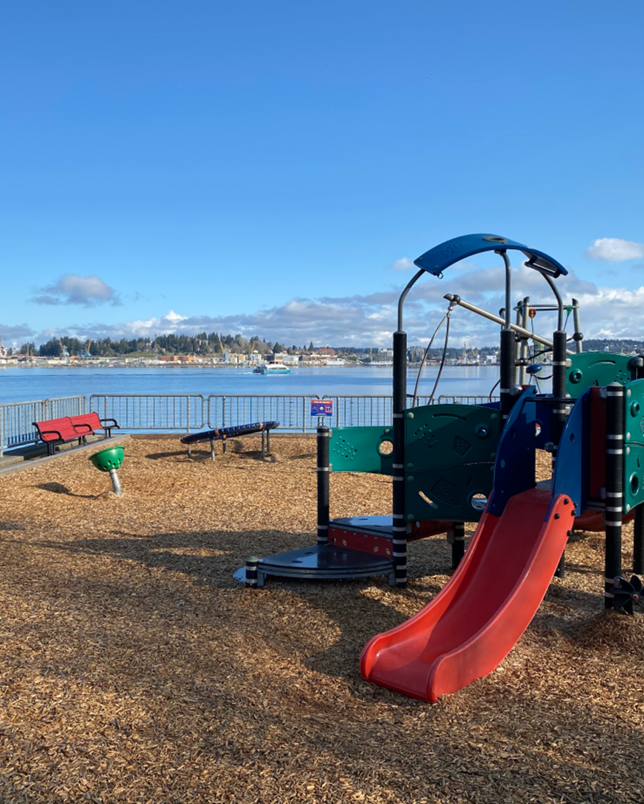 A park by a body of water with a red slide attached to a playset