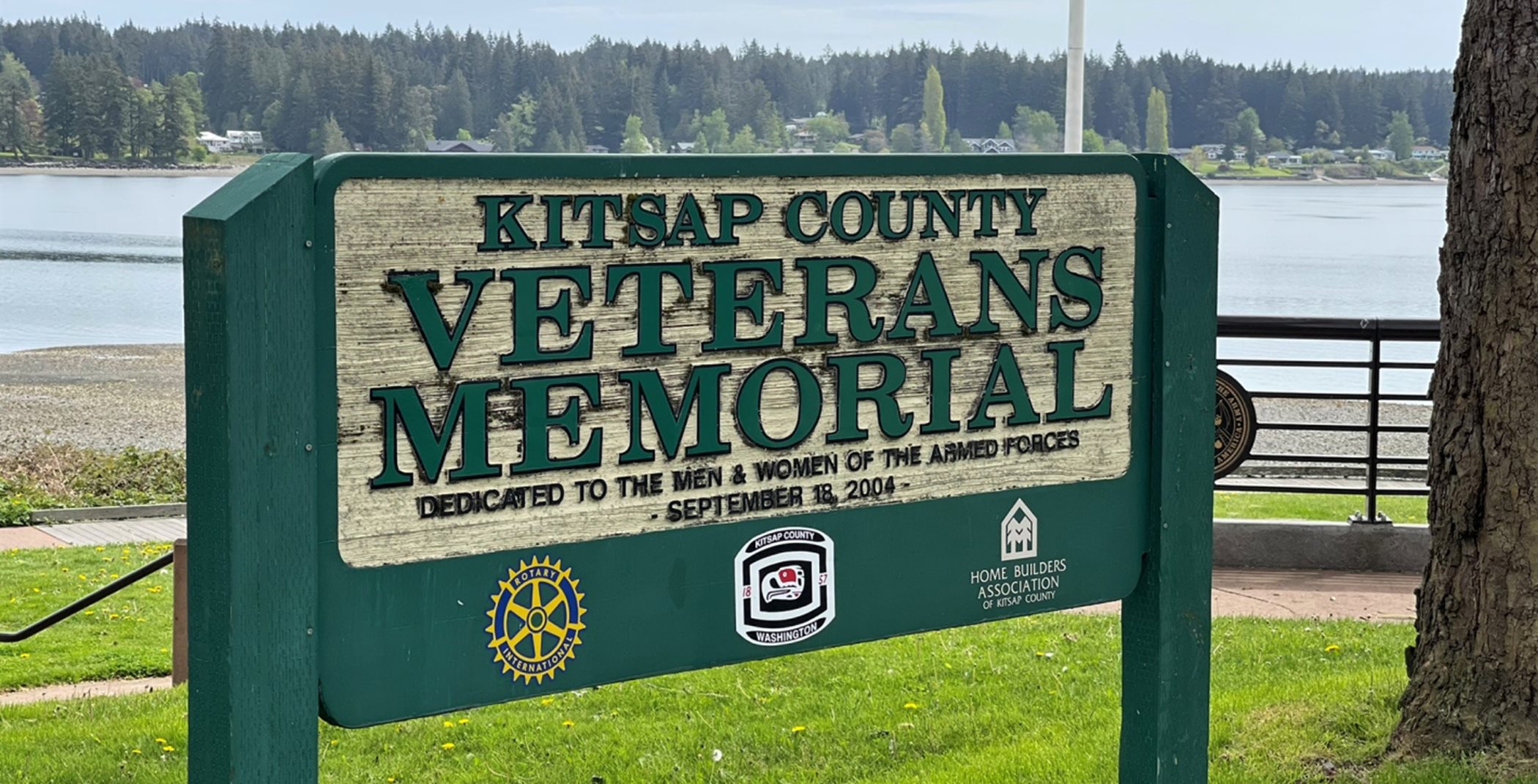 Veteran's Memorial Park sign. It is green and yellow. It says: "Kitsap County Veterans' Memorial. Dedicated to the Men and Women of the Armed Forces. September 18, 2004"