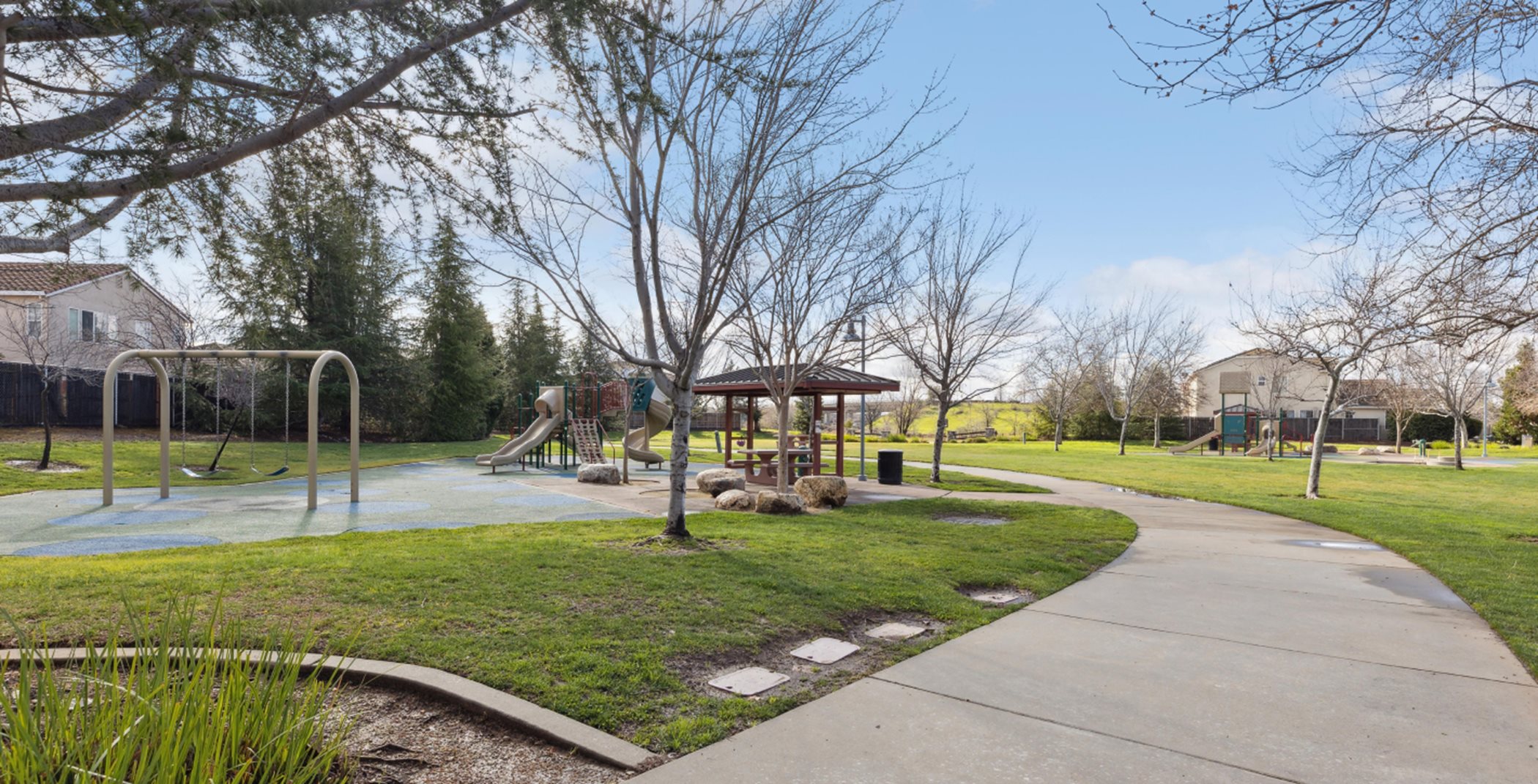 Paved path through the park surrounded by grass, trees, and a swingset