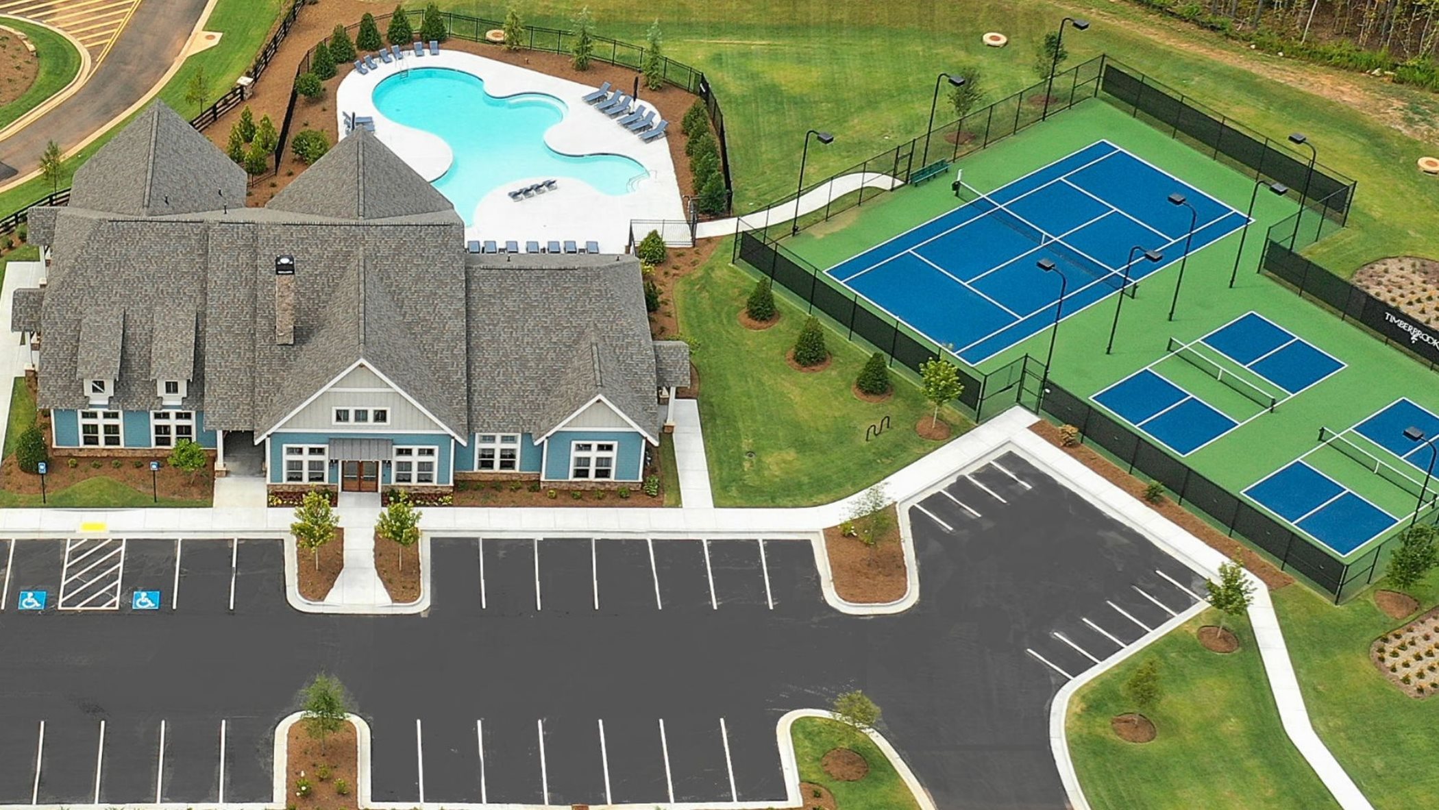A blue, white, and grey clubhouse in front of a spacious parking lot. In the backyard there is a whimsically shaped pool and sport court with tennis and pickle ball.